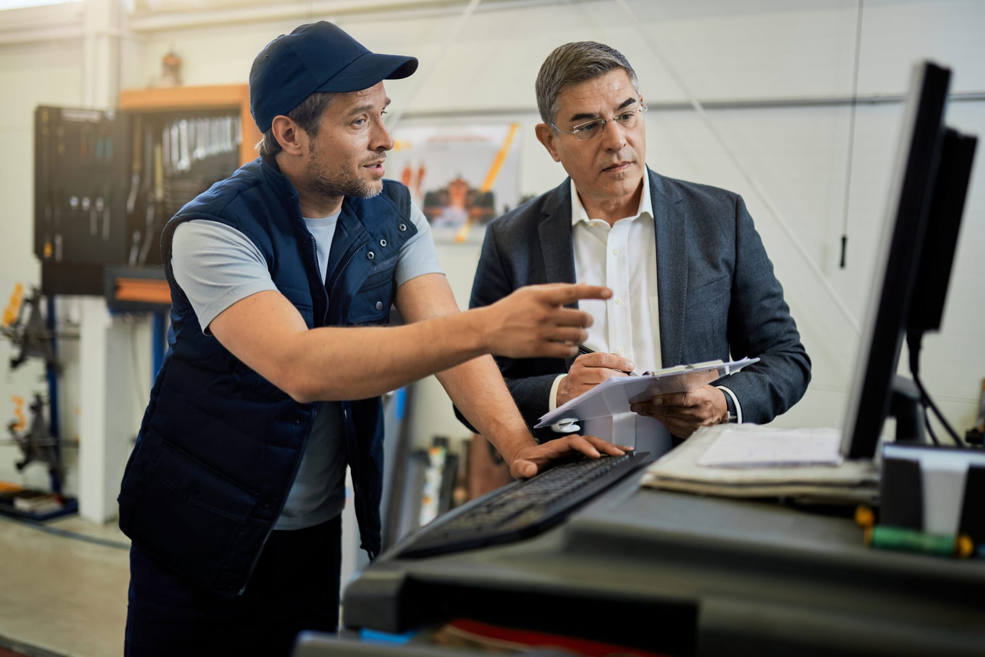 Mid adult manager and car mechanic talking while using computer in auto repair shop.